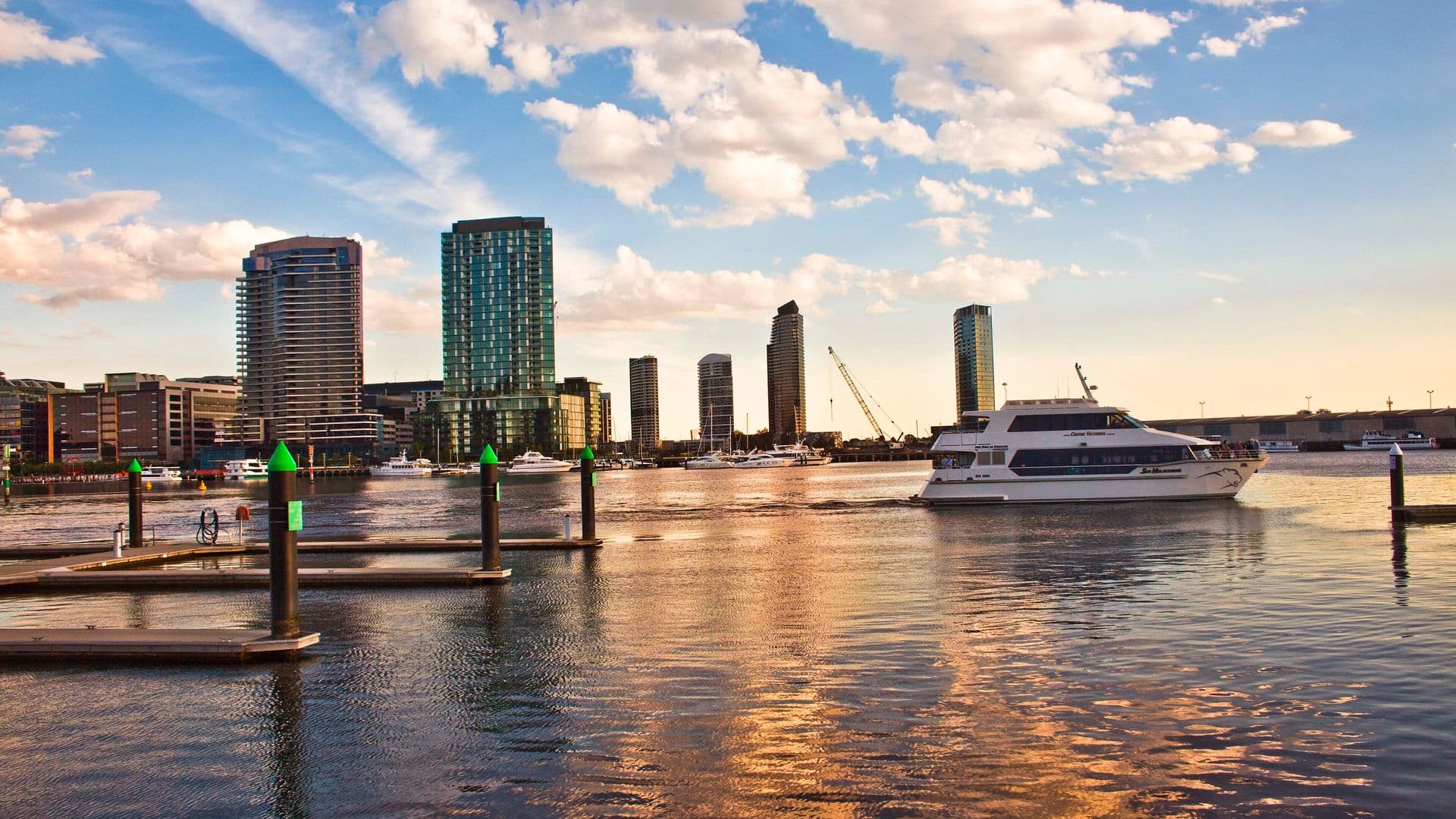 Jahta u plovidbi kraj Docklandsa u Melbourneu, s neboderima i pontonima u prvom planu