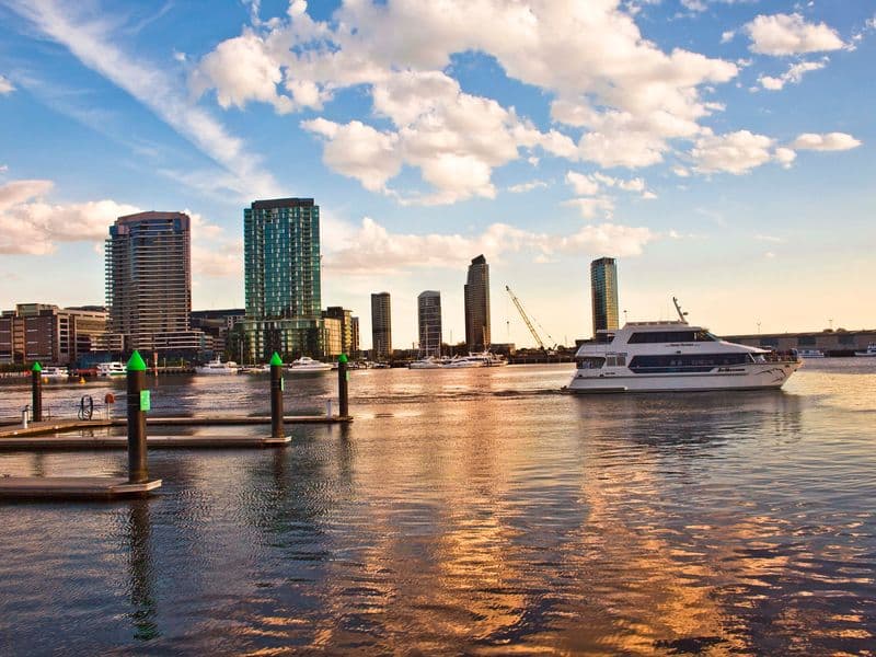 Jahta u plovidbi kraj Docklandsa u Melbourneu, s neboderima i pontonima u prvom planu